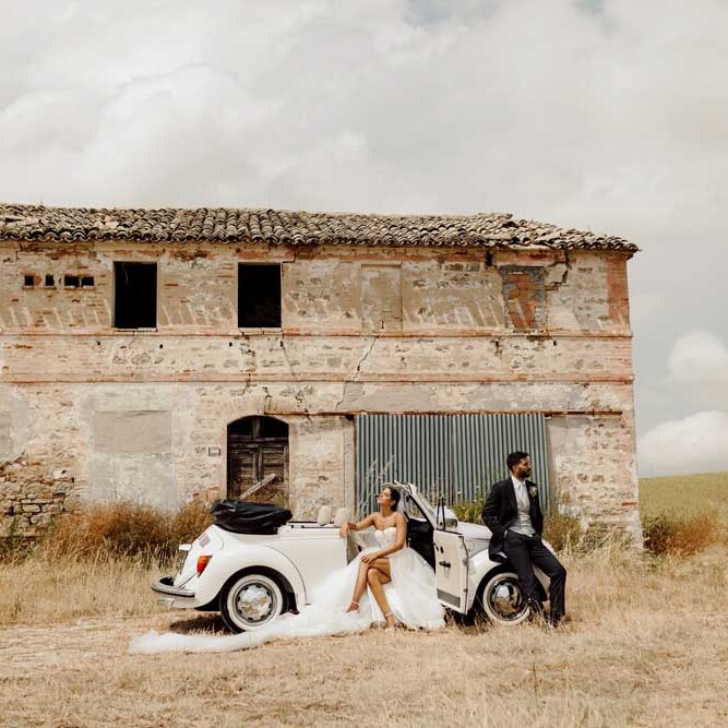 Sposi seduti su Maggiolino Volkswagen bianco vintage davanti a edificio rustico, matrimonio a Jesi nelle Marche
