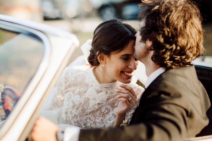 Sposo che bacia la tempia della sposa in auto d'epoca, lei sorride a occhi chiusi, abito da sposa in pizzo, luce calda dorata, momento intimo matrimonio Marche
