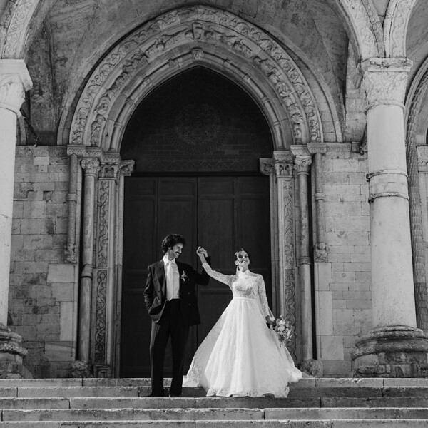 Sposi che si tengono per mano sulle scale del Duomo di Osimo, lei in abito da sposa con maniche in pizzo, lui in abito scuro, portale romanico sullo sfondo, foto in bianco e nero